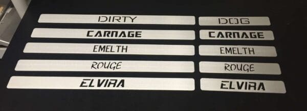 Metal plates with word cutouts: DIRTY DOG, CARNAGE, EMELTH, ROUGE, ELVIRA, displayed on a black surface.