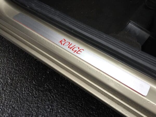 ROUGE engraved on a car door sill, with a metallic finish and a black interior visible.
