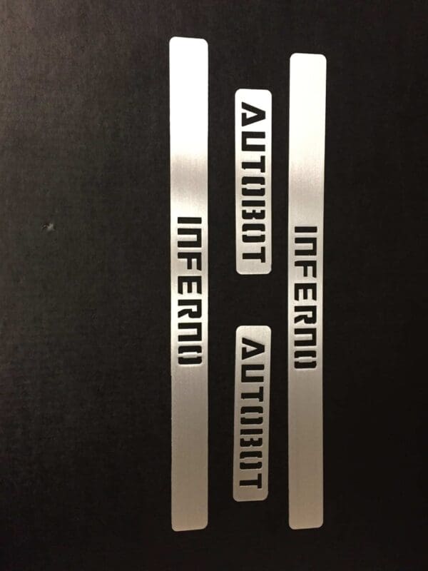 Silver metal pieces with cutouts spelling "AUTOBOT" and "INFERNO" arranged vertically on a black background.