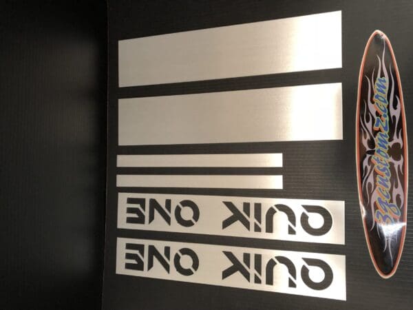 Metal pieces with cutouts of the word "ONE" and a decorative design labeled "3genstomz.com" on a dark background.