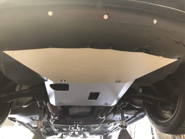 Underside of a vehicle showing a metal skid plate secured with bolts, with visible wheels and suspension components.