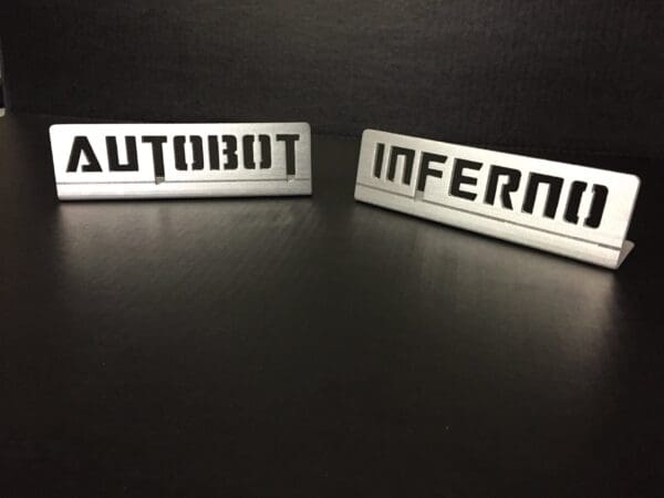 Two metal pieces with cut-out text: "AUTOBOT" on the left and "INFERNO" on the right, set against a dark background.