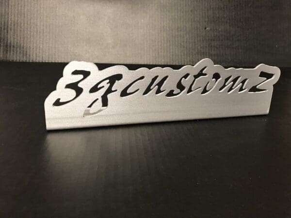Silver metal nameplate with "3Fcustomz" cut out, placed on a black table.