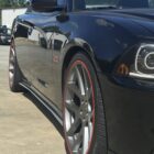 Black car with silver wheels and red accents parked on concrete, reflecting nearby trees and clouds.