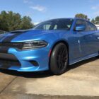 A blue Dodge Charger with black racing stripes parked on a concrete surface under a clear sky.