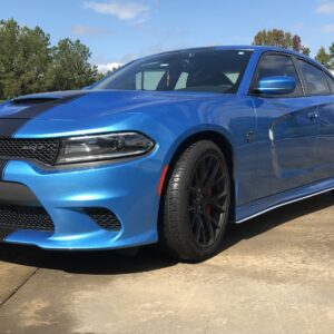 A blue Dodge Charger with black racing stripes parked on a concrete surface, surrounded by trees.