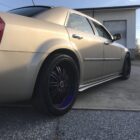 A gold sedan with custom black wheels parked on a gravel surface near a building.
