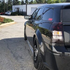 Black car parked on a paved lot, with a building and trees in the background.