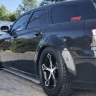 A black car with custom wheels and a sticker parked on a paved lot, surrounded by greenery and buildings.
