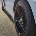 Close-up of a sleek car's black wheel and tire on a paved surface, sunlight reflecting off the vehicle's side.
