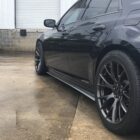 A black car with large wheels parked on a wet concrete surface near a building.