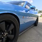 A blue muscle car with black wheels parked on a concrete surface, reflecting the sky and nearby buildings.