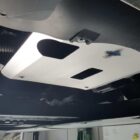 A metallic skid plate is mounted under a vehicle, with visible cutouts and a sticker.