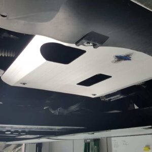A metallic skid plate is mounted under a vehicle, with visible cutouts and a sticker.