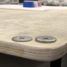 Two metal washers sit on the edge of a wooden surface, with a blurred background of tools and materials.