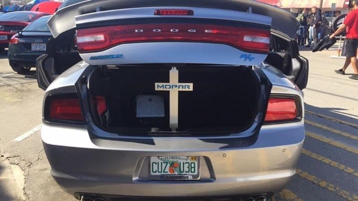 Open trunk of a silver Dodge car displaying a Mopar cross and a license plate. People are visible in the background.