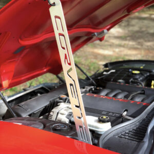 A red car hood is open, revealing a Corvette engine and a supporting prop with "CORVETTE" engraved.