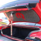 A red car trunk is open, featuring a decorative emblem and a strap for support.
