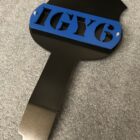Black and blue sign with "IGY6" cut out, resting on a textured surface.