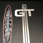 A silver "GT" emblem and a colorful surfboard sticker on a black background.