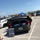 A black car trunk is open, displaying items inside, with a sign nearby detailing the vehicle's features.